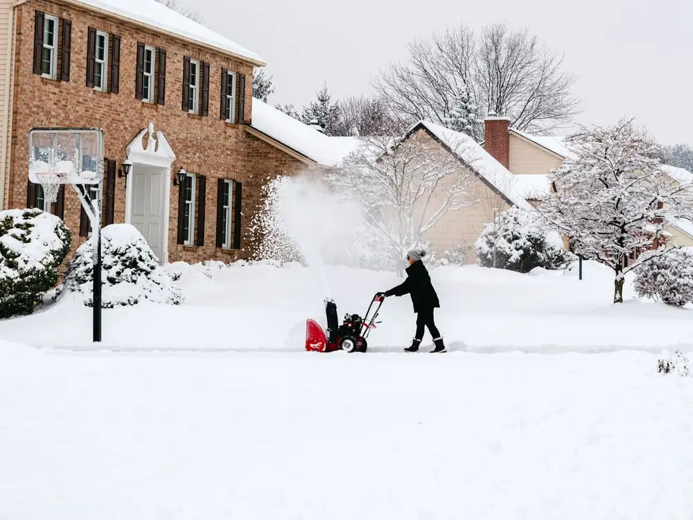 A snow blower clearing a driveway in front of a brick house after a heavy snowfall